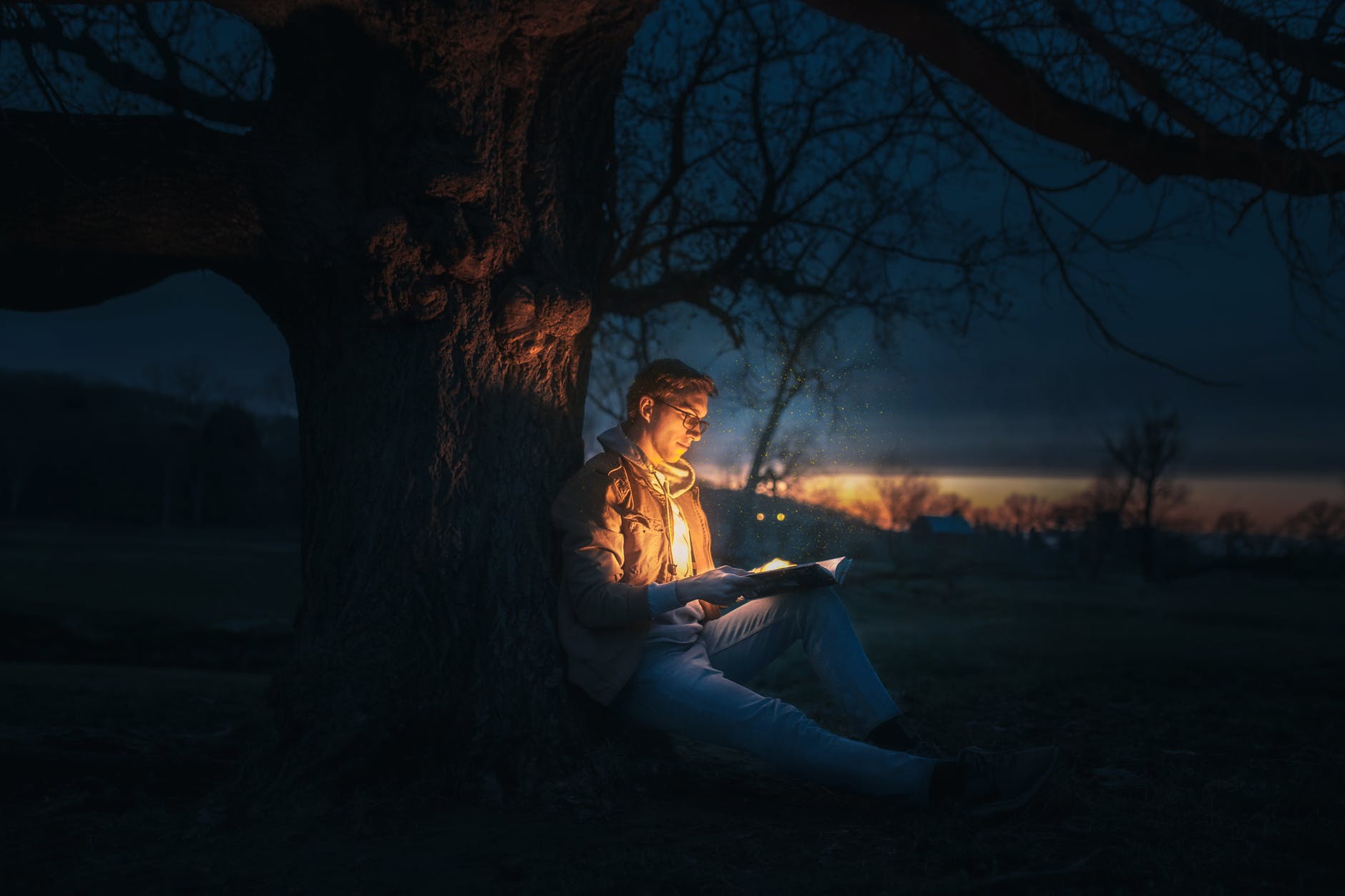 man sitting under a tree reading a book during night time